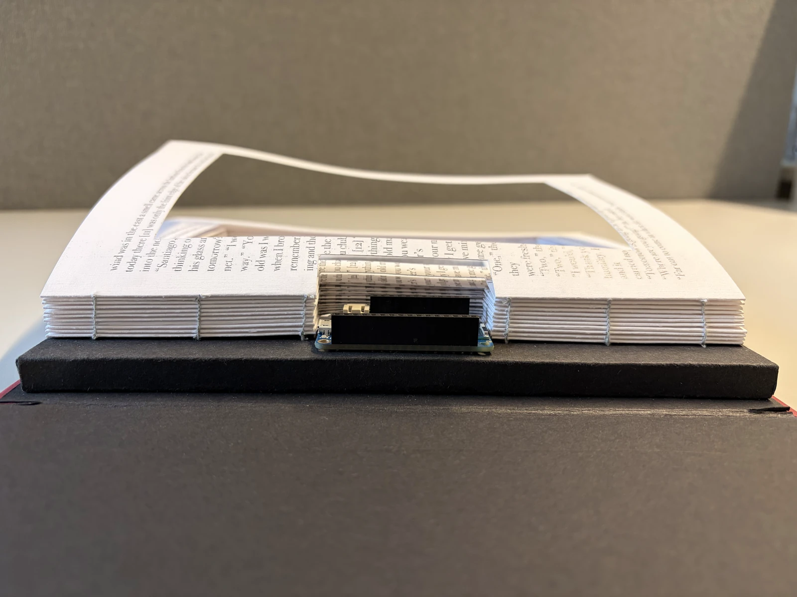 The book seen from its foot: the hand-bound page block over the dark cover, with the Arduino Feather at the spine