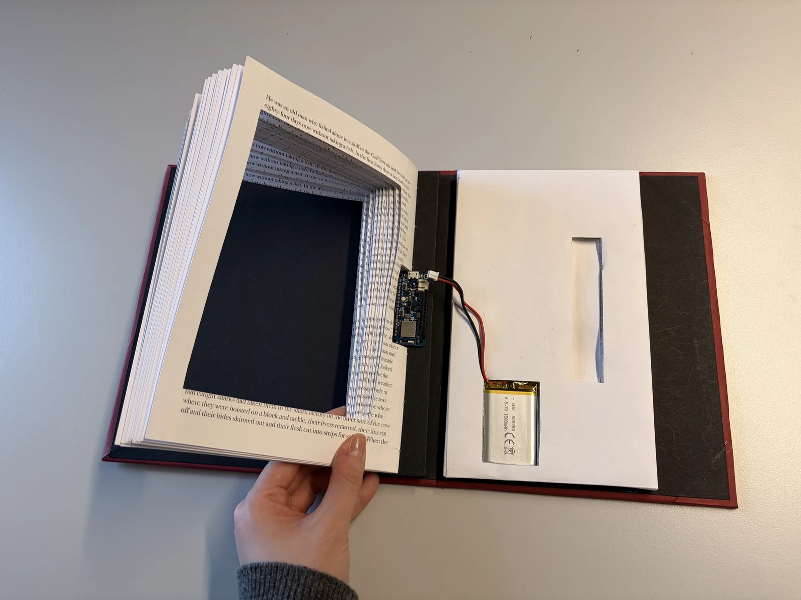 The pages lifted to show the hollow cut out of them, the Arduino Feather and the battery set into the cover